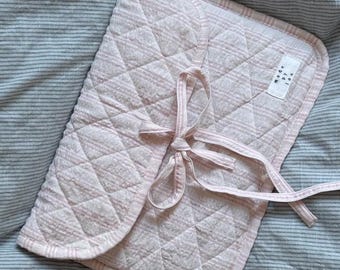 May include: A pink quilted baby changing pad with a white and pink striped pattern and a white tag that says "Made by Joy" in black text.