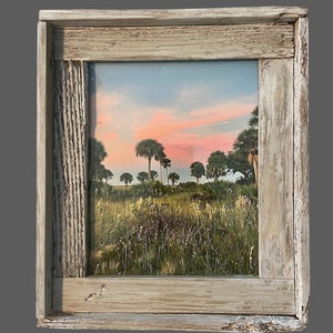 May include: A rustic wooden frame with a faded white finish encloses a photograph of a field of tall grass with palm trees in the background. The sky is a soft pink and orange.