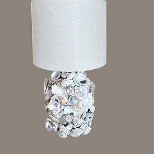 May include: A decorative table lamp with a cylindrical, off-white linen lampshade. The base is crafted from a cluster of white and gray seashells. The lamp is a unique home decor item.