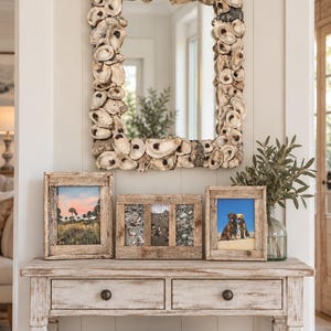 May include: A rectangular wall mirror framed with numerous overlapping oyster shells in shades of cream, gray, and brown. The mirror is positioned above a wooden console table featuring three rustic picture frames and a glass vase with greenery.