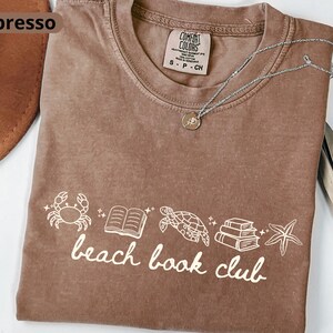 May include: An espresso-colored t-shirt featuring a white graphic print of a crab, an open book, a sea turtle, a stack of books, and a starfish, with the text "beach book club" written below the icons in a cursive font.