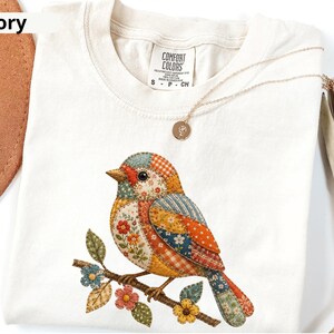 May include: An ivory-colored t-shirt featuring a graphic of a bird perched on a branch. The bird is composed of various patchwork patterns in shades of orange, blue, red, and yellow. The shirt has a Comfort Colors label at the neck and a gold necklace.