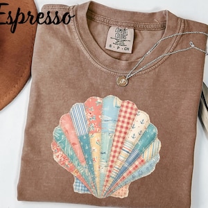 May include: Espresso-colored t-shirt with a seashell design made of patchwork fabric. The shell features various patterns, including floral, striped, and gingham. A silver necklace with a small pendant rests on the shirt. The shirt has a Comfort Colors tag.