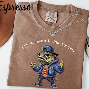 May include: Espresso-colored t-shirt with the phrase "Off to commit dad foolery" and a cartoon fish wearing sunglasses, a backwards cap, and a gold chain. The shirt has a crew neck and short sleeves. A silver necklace is draped across the shirt.