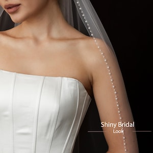 May include: A strapless white satin wedding dress with vertical seams. A sheer veil with a pearl trim cascades down the arm. The text "Shiny Bridal Look" is visible.