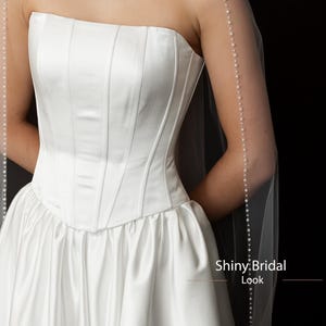 May include: A white strapless bridal gown with a fitted bodice and a full skirt. A sheer veil with a pearl trim drapes over the shoulders. The text "Shiny Bridal Look" is visible.