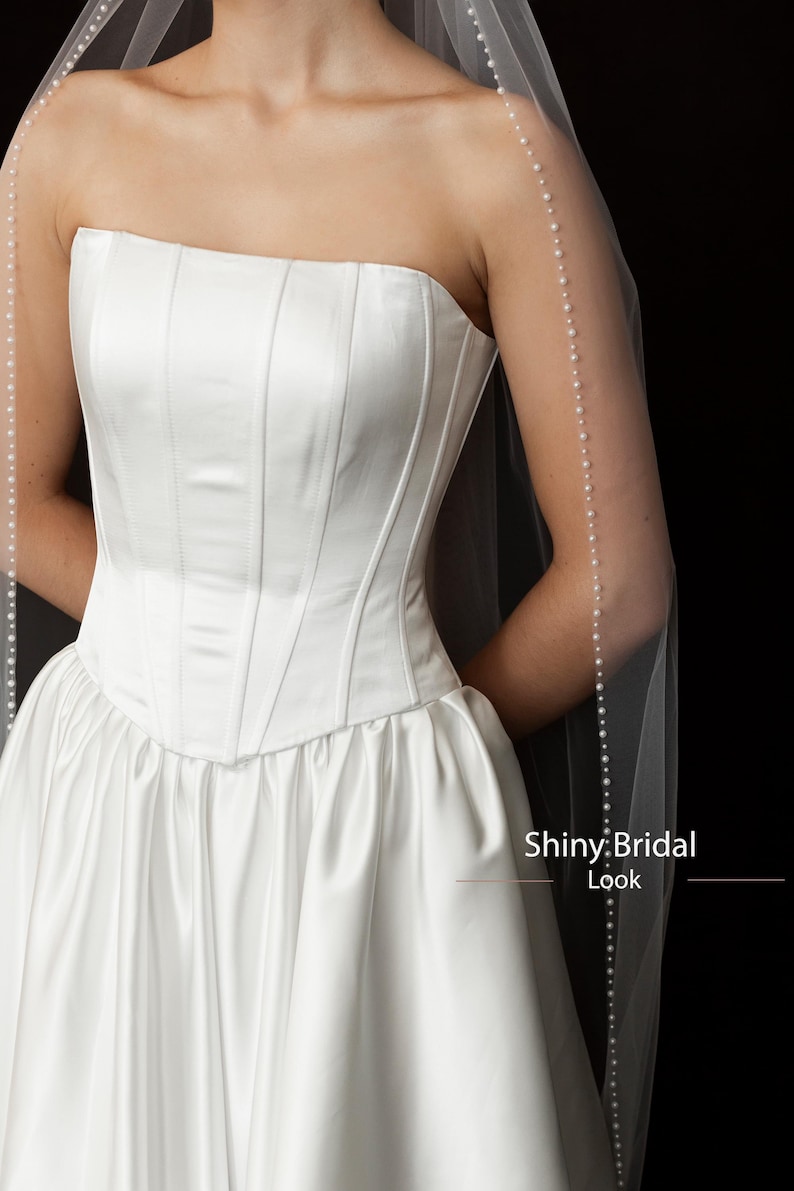 May include: A white strapless bridal gown with a fitted bodice and a full skirt. A sheer veil with a pearl trim drapes over the shoulders. The text "Shiny Bridal Look" is visible.