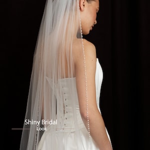 May include: A white bridal veil with a beaded edge and a long, flowing design. The veil is paired with a white wedding dress. The image has the text "Shiny Bridal Look".