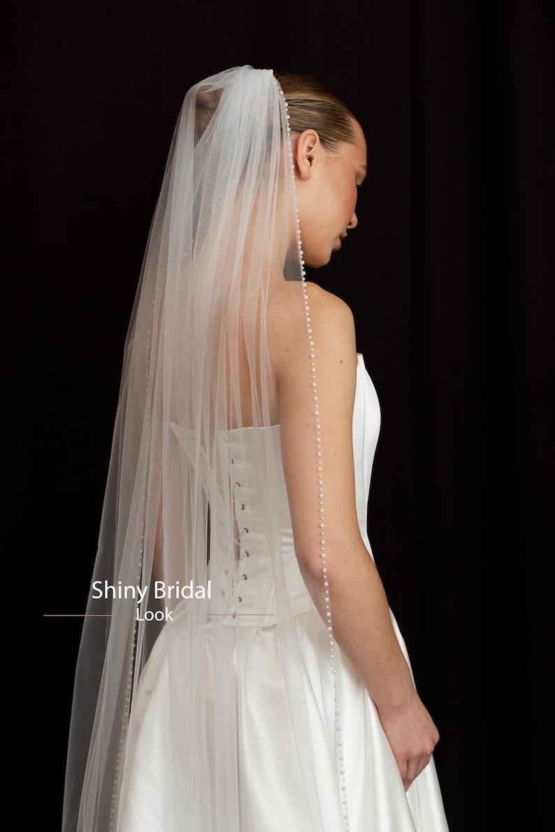 May include: A white bridal veil with a beaded edge and a long, flowing design. The veil is paired with a white wedding dress. The image has the text "Shiny Bridal Look".
