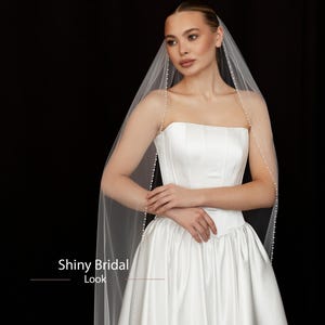 May include: A white bridal gown with a strapless bodice and a full skirt. A sheer veil, adorned with small pearls, drapes over the shoulders. The text "Shiny Bridal Look" is visible in the image.