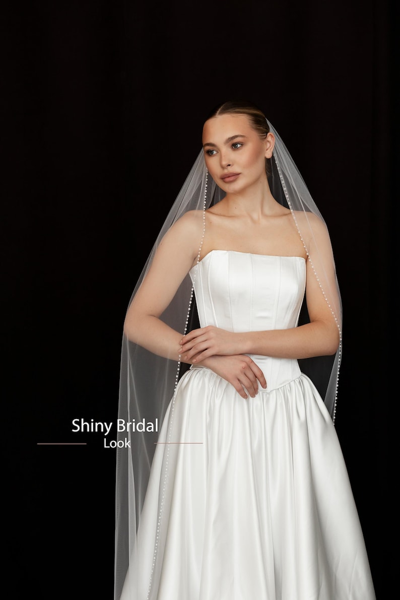 May include: A white bridal gown with a strapless bodice and a full skirt. A sheer veil, adorned with small pearls, drapes over the shoulders. The text "Shiny Bridal Look" is visible in the image.