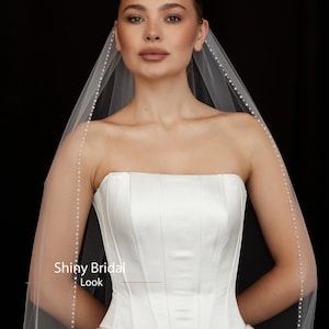 May include: A strapless white satin wedding dress with a structured bodice and a flowing skirt. A sheer veil with pearl accents drapes over the shoulders. The text "Shiny Bridal Look" is visible.