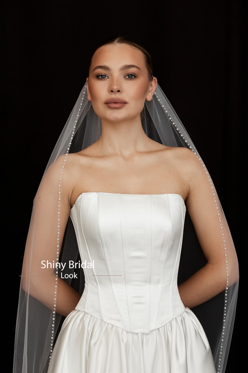 May include: A strapless white satin wedding dress with a structured bodice and a flowing skirt. A sheer veil with pearl accents drapes over the shoulders. The text "Shiny Bridal Look" is visible.