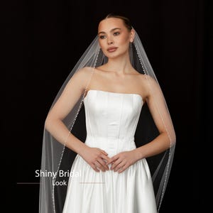 May include: A strapless white satin wedding dress with a full skirt. A sheer veil, edged with small pearls, drapes over the shoulders. The text "Shiny Bridal Look" is visible at the bottom of the image. The background is black.