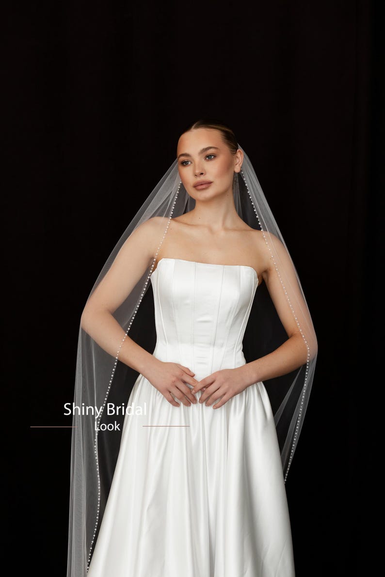 May include: A strapless white satin wedding dress with a full skirt. A sheer veil, edged with small pearls, drapes over the shoulders. The text "Shiny Bridal Look" is visible at the bottom of the image. The background is black.