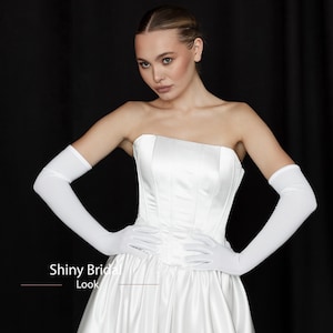 Op de afbeelding: Witte satijnen trouwjurk met een strapless ontwerp en een nauwsluitend lijfje. Het model draagt lange witte handschoenen. De tekst "Shiny Bridal Look" is zichtbaar in de afbeelding.