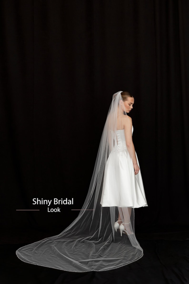 May include: A white bridal dress with a short skirt and a long, sheer veil. The veil is trimmed with small beads. The text "Shiny Bridal Look" is visible in the image. The background is black.