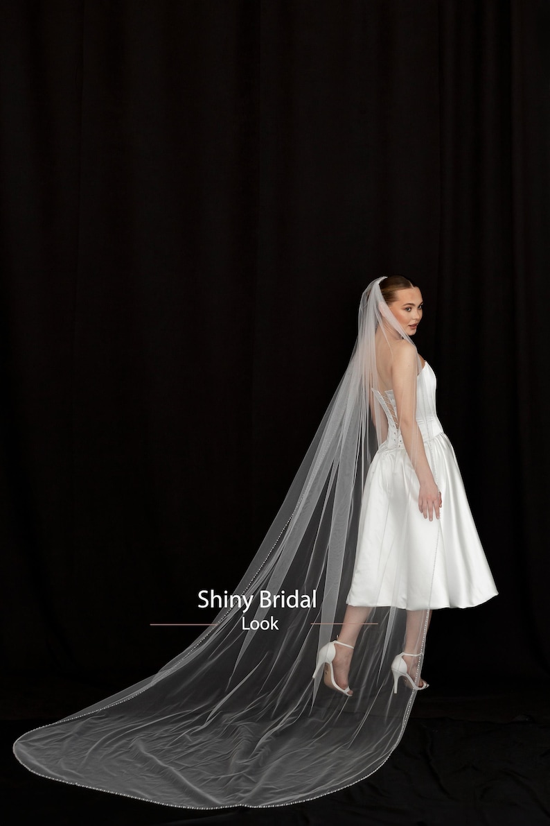 May include: A bride in a white satin wedding dress and long veil. The veil is trimmed with small beads and trails behind the bride. The text "Shiny Bridal Look" is visible in the image.