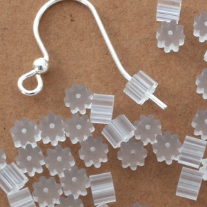 May include: A collection of clear, translucent earring backs and a silver-colored earring hook. The earring backs are small, with a flower-like design and a rectangular shape. The hook is curved with a small ball at the end.