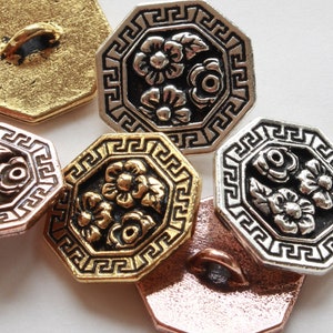 May include: Close-up of several decorative metal buttons in various colors, including gold, silver, and copper. Each button has an octagonal shape with a floral design in the center and a Greek key border.