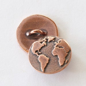 May include: Two antique copper-colored buttons. One button is face up, showing the back with a loop. The other button is face down, displaying a detailed world map design. The buttons are round and have a vintage aesthetic.