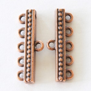 May include: Two copper-colored multi-strand jewelry clasps. Each clasp has four loops and a rectangular bar with a dotted design down the center. The clasps are designed to connect multiple strands of beads or chains.