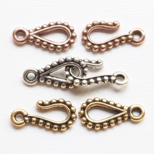 May include: An assortment of jewelry clasps in various metallic finishes. The clasps feature a teardrop shape with a beaded border and a hook-and-eye closure. Finishes include copper, silver, and gold tones. These clasps are ideal for handmade jewelry.