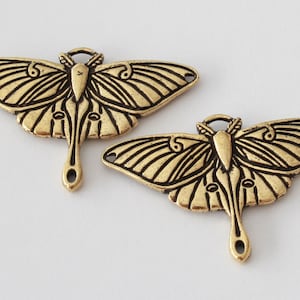 May include: Two antique gold-colored moth-shaped pendants. Each pendant features detailed wing patterns, a central body, and a loop for hanging. The pendants have a vintage, aged appearance, suggesting they are made of brass or a similar metal.