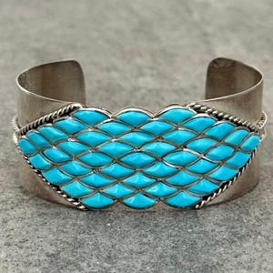May include: A silver cuff bracelet with a central turquoise inlay. The turquoise stones are arranged in a repeating pattern, framed by a twisted silver rope design. The bracelet has a polished finish.