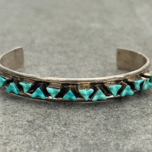 May include: A silver cuff bracelet with turquoise triangular stones. The bracelet has a textured band with a row of turquoise stones set in a repeating pattern. The bracelet is open at the ends.