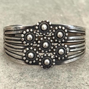 May include: A silver cuff bracelet with a floral design. The bracelet features a series of parallel bands with etched lines and a central floral cluster of beaded accents. The bracelet has a vintage, handcrafted appearance.