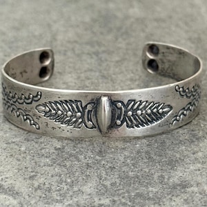 May include: A silver cuff bracelet with an engraved design. The bracelet features a central oval shape flanked by detailed leaf and scroll patterns. The open ends have two small holes each.