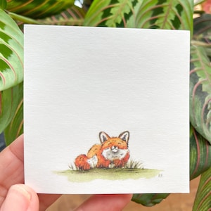 May include: A watercolor painting of a small, orange fox sitting on a green patch of grass. The fox has a white chest and a black nose.
