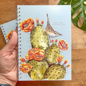 May include: A blue spiral bound notebook with a watercolor illustration of a cactus wren perched on a prickly pear cactus with orange flowers. The text "Cactus Wren Campylohynchus brunneicapillus" is printed below the bird.
