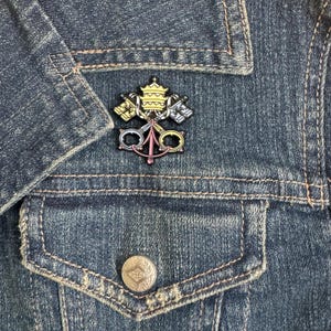 May include: A gold, silver, and pink enamel pin featuring the Papal Keys, a symbol of the papacy. The pin is attached to a denim jacket.