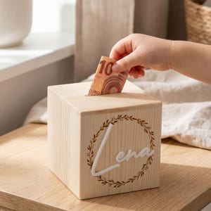 Könnte beinhalten: Eine hellfarbene Holzspardose in Würfelform. Die Box hat oben einen Schlitz zum Einwerfen von Geld. Auf der Vorderseite der Box befindet sich der Name "Lena" in weißer Schrift, umgeben von einem dekorativen Kranz.