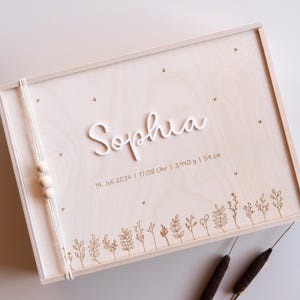 Könnte beinhalten: Eine Holz-Erinnerungskiste mit einer weißen Aufschrift, die "Sophia" und das Datum "14. Juli 2024 | 17:08 Uhr | 3,940 g | 54 cm" lautet. Die Kiste ist mit einer Reihe kleiner, gravierter Blumenmuster verziert.