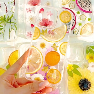 May include: Transparent phone cases decorated with dried flowers and fruit. Designs include pink flowers, yellow sunflowers, and orange citrus slices. The cases are held by a hand, showcasing the variety of styles.