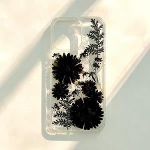 May include: Clear phone case with pressed black flowers and fern-like leaves. The case has a rectangular shape with rounded corners and cutouts for the camera and buttons. The floral design is visible against a white background.