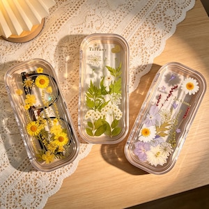 Puede incluir: Tres estuches rectangulares transparentes para gafas con diseños florales. Un estuche contiene flores amarillas y gafas. Los otros dos estuches presentan flores prensadas y vegetación. Un estuche está etiquetado "Tiffany" y otro "Jackson."