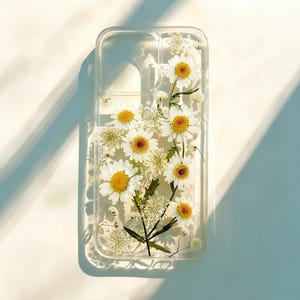 May include: Clear phone case with embedded white daisies and yellow centers. The case has a rectangular shape with rounded corners. The flowers are arranged in a natural, organic pattern, with green stems and leaves.