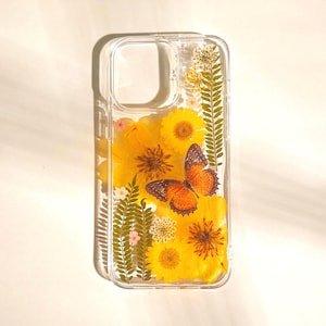 May include: Clear phone case with pressed yellow flowers and a butterfly. The case features a transparent design, showcasing the natural beauty of the floral arrangement and the orange butterfly. A unique and artistic accessory.