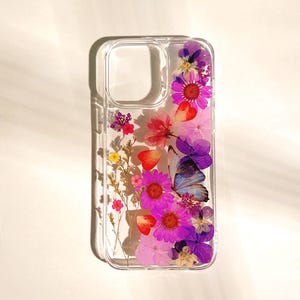 May include: Clear phone case with pressed flowers and a butterfly. The case features a variety of colorful flowers in shades of pink, purple, and red. A blue and purple butterfly is also embedded in the design.