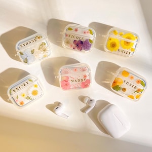 May include: Clear protective cases for wireless earbuds, each adorned with floral designs and personalized names: Yvonne, Nicole, Emily, Kelsey, Maddy, and Susie. Includes a charging case and earbuds.