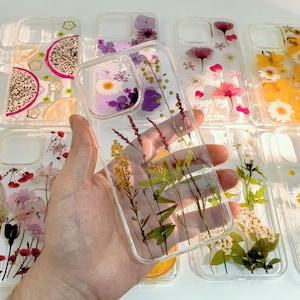 May include: Clear phone cases with dried flowers and fruit slices embedded in resin. The cases feature various floral arrangements with yellow, purple, and pink flowers. Some cases have dragon fruit and lemon slices.
