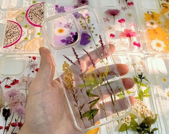 May include: Clear phone cases with dried flowers and fruit slices embedded in resin. The cases feature various floral arrangements with yellow, purple, and pink flowers. Some cases have dragon fruit and lemon slices.