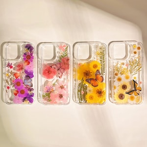 May include: Four clear phone cases with floral and butterfly designs. The cases feature pressed flowers in shades of pink, purple, yellow, and white. Butterflies in orange and purple add a decorative touch to the cases.