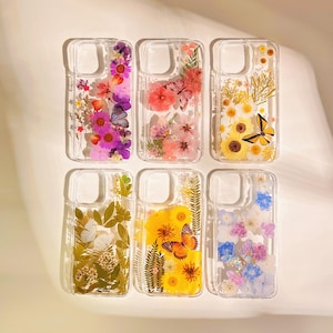 May include: Six clear phone cases with floral and butterfly designs. Each case features a unique arrangement of dried flowers and butterflies in various colors, including pink, yellow, and purple. The cases are arranged in two rows of three.