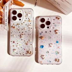 May include: Two clear phone cases with glitter and small decorative charms. The case on the left has a gold glitter background with white and gold charms. The case on the right has a pink glitter background with white and blue charms. The case on the left has the name "Amelia" spelled out in gold letters. The case on the right has the name "Lilia" spelled out in gold letters.