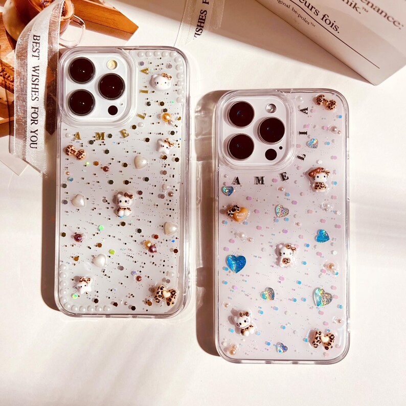 May include: Two clear phone cases with glitter and small decorative charms. The case on the left has a gold glitter background with white and gold charms. The case on the right has a pink glitter background with white and blue charms. The case on the left has the name "Amelia" spelled out in gold letters. The case on the right has the name "Lilia" spelled out in gold letters.